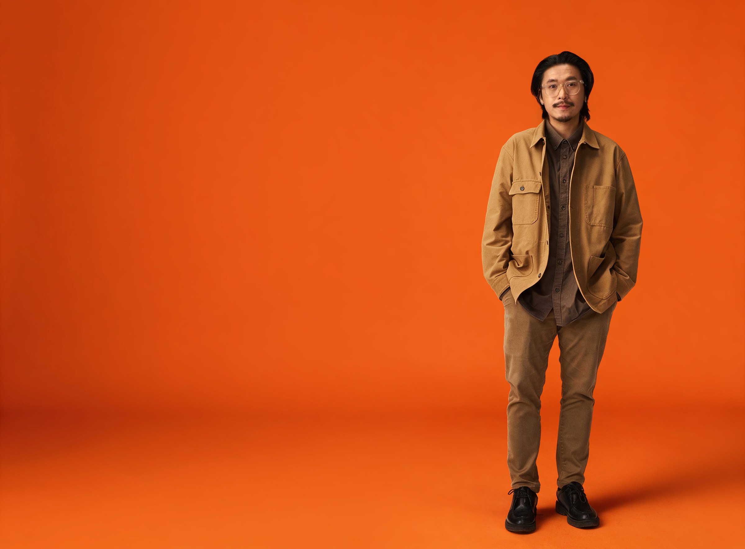Baz Poonpiriya standing in a tan work jacket against a dark green backdrop