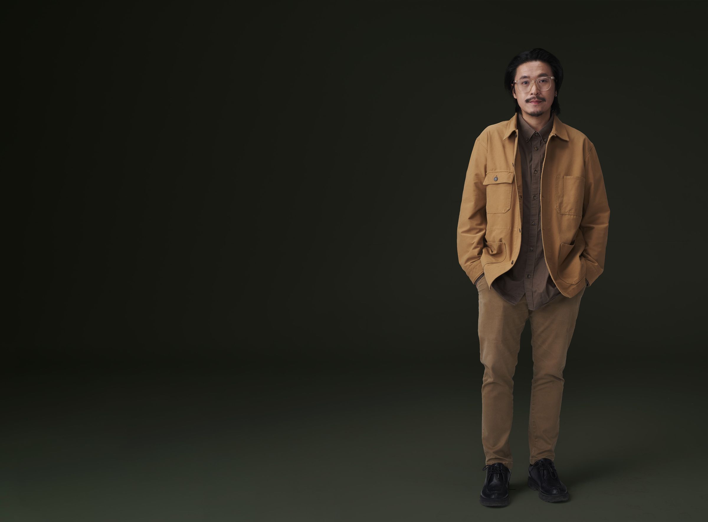 Baz Poonpiriya standing in a tan work jacket against a dark green backdrop
